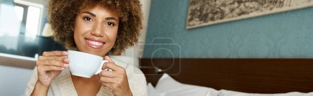 cheerful young african american woman with curly hair savoring cappuccino in hotel room, banner