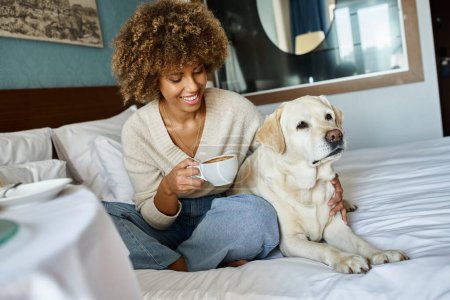 Téléchargez les photos : Femme afro-américaine gaie tenant tasse et câlinant son chien labrador dans la chambre d'hôtel acceptant les animaux - en image libre de droit