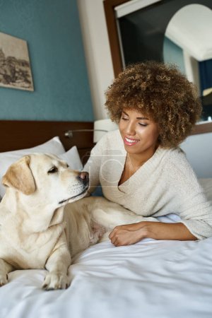 fröhliche afrikanisch-amerikanische Frau liegt auf dem Bett und kuschelt ihren niedlichen Labrador in haustierfreundlichem Hotel