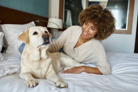 smiling african american woman lying on bed and cuddling her labrador dog in pet friendly hotel