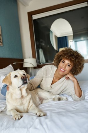 young african american woman lying on bed and cuddling her labrador dog in pet friendly hotel