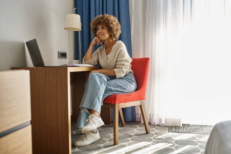 mujer afroamericana joven soñadora trabajando a distancia en la habitación del hotel, usando el ordenador portátil y tomando notas