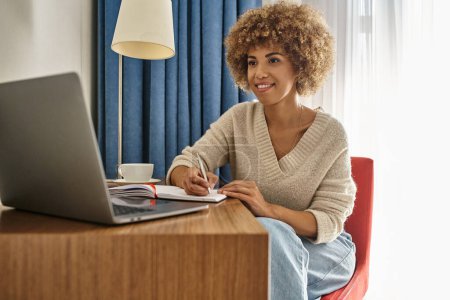 mujer afroamericana feliz a distancia trabajando en la habitación del hotel, usando el ordenador portátil y tomando notas