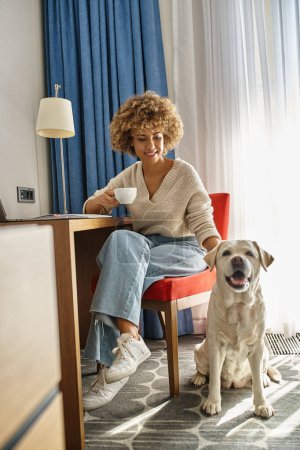 mujer afroamericana disfruta del café y trabaja remotamente cerca de su labrador en un hotel que acepta mascotas