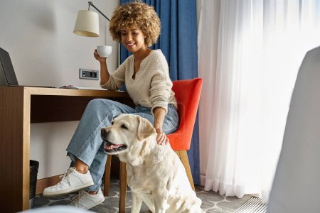 mujer afroamericana feliz disfruta del café y trabaja cerca de su labrador en un hotel que acepta mascotas