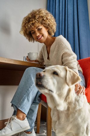 Happy and curly african american woman enjoys coffee near labrador in a pet-friendly hotel