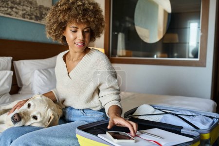 Glückliche Afroamerikanerin sitzt mit ihrem Labrador in der Nähe von geöffnetem Gepäck und greift zum Smartphone