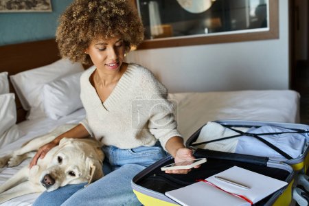 Happy african american woman sitting with her labrador near open luggage and checking her smartphone