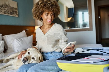 Happy african american woman sitting with her labrador near open luggage and using smartphone