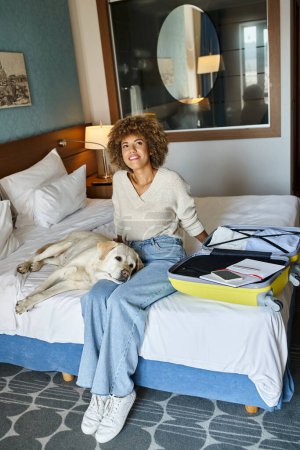 femme afro-américaine joyeuse assise avec chien labrador près des bagages ouverts dans un hôtel acceptant les animaux de compagnie