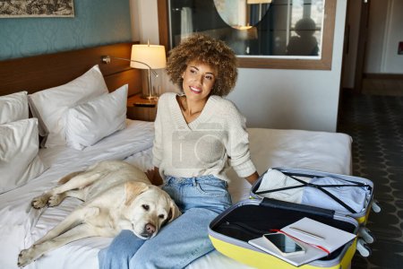 young african american woman sitting with labrador dog near open luggage in a pet-friendly hotel