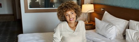 Smiling  and dreamy African American woman with curly hair sitting on hotel bed, travel banner