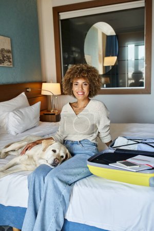 jolly african american woman sitting with labrador dog near open luggage in a pet-friendly hotel