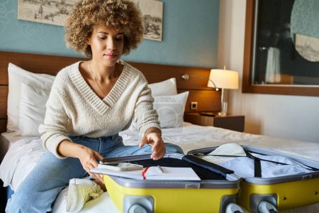 young African American woman with curly hair unpacking her luggage in hotel room, getaway concept
