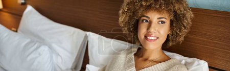 femme afro-américaine rêveuse et heureuse se détendre sur un lit d'hôtel et regarder ailleurs, bannière