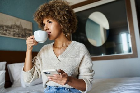 mujer afroamericana de pelo rizado bebiendo café mientras usa el teléfono inteligente en la habitación de hotel, viajes