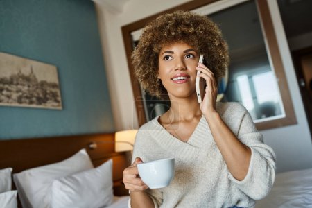 happy african american woman holding cup of coffee while having phone call in hotel room