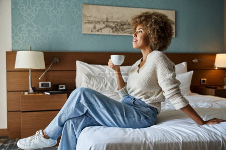 mujer afroamericana feliz y joven disfrutando del café mientras está sentada en la cama del hotel, escapada de fin de semana