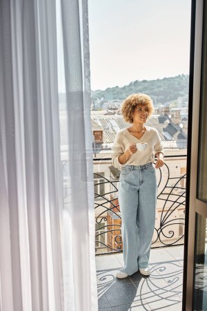 fröhliche lockige afrikanisch-amerikanische Frau steht mit Tasse Kaffee auf Hotelbalkon, Blick auf die Stadt