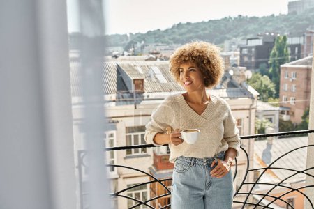 dreamy curly african american woman standing with cup of coffee on hotel balcony, city view