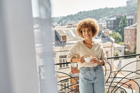 happy curly african american woman standing with cup of coffee on hotel balcony, city view