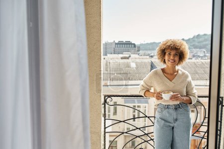 smiling curly african american woman standing with cup of coffee on hotel balcony, city view