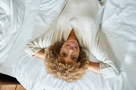 top view of relaxed african american woman smiling and lying on bed in hotel room, upside down