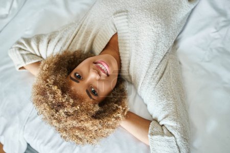 top view of happy african american woman smiling and lying on bed in hotel room, upside down