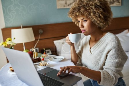 curly african american woman using her laptop near breakfast in hotel, room service and convenience