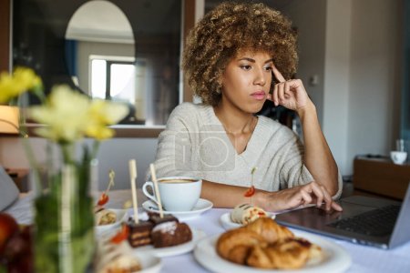pensive african american woman using laptop near breakfast in hotel, room service and convenience