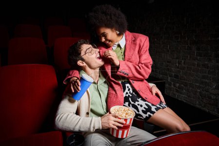 fröhliche afrikanisch-amerikanische Frau teilt Popcorn mit ihrem Freund, während sie auf seinem Schoß sitzt