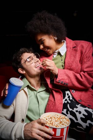 fröhliches multiethnisches Paar im Retro-Gewand teilt Popcorn bei einem Date am Valentinstag im Kino