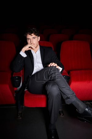 handsome man in black suit sitting on red cinema chair and looking at camera with hand near face