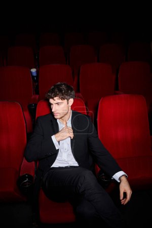 attractive stylish male model in black elegant suit sitting on red chair at cinema and looking away