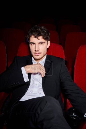 good looking elegant male model in black chic suit sitting on red cinema chair and looking at camera
