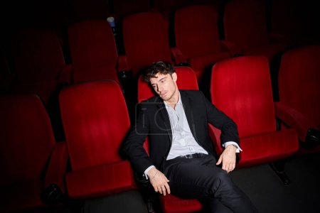 beau modèle masculin élégant en costume élégant noir assis sur une chaise de cinéma rouge et regardant ailleurs