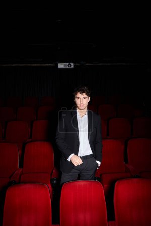young appealing man in elegant black suit posing near red cinema chairs and looking at camera