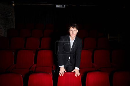 good looking young man in black elegant suit posing between red cinema chairs and looking at camera