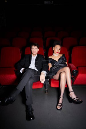 junges multiethnisches, elegantes Paar in schwarzen Anzügen, das an ihrem Date Zeit miteinander im Kino verbringt