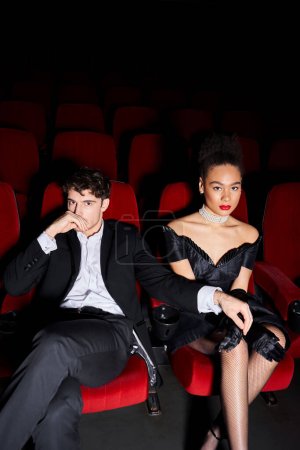 Reizvolles multikulturelles Paar posiert gemeinsam auf roten Kinosesseln und schaut beim Date in die Kamera