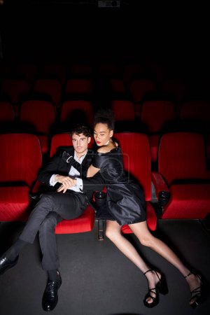 Téléchargez les photos : Attrayant beau couple diversifié étreignant sur des chaises de cinéma rouge à la date de la Saint-Valentin - en image libre de droit