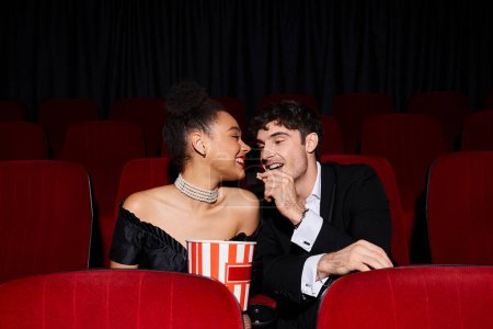 Téléchargez les photos : Beau homme partageant pop-corn avec sa jeune copine afro-américaine sur la date au cinéma - en image libre de droit