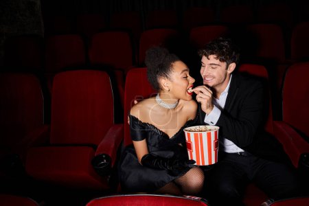 joyful handsome man in black suit feeding popcorn to his african american young girlfriend on date