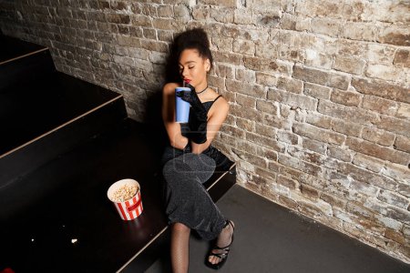 Téléchargez les photos : Séduisante femme afro-américaine élégante en robe noire avec rouge à lèvres posant avec boisson au cinéma - en image libre de droit