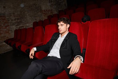 Téléchargez les photos : Attrayant jeune modèle masculin en costume noir élégant assis sur une chaise rouge au cinéma et regardant loin - en image libre de droit