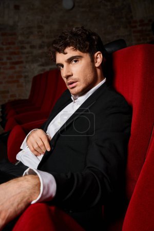 Téléchargez les photos : Séduisant bel homme en élégant costume noir assis sur une chaise de cinéma rouge et regardant la caméra - en image libre de droit