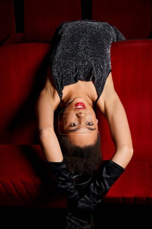 alluring young african american female model lying on red cinema chairs and looking at camera