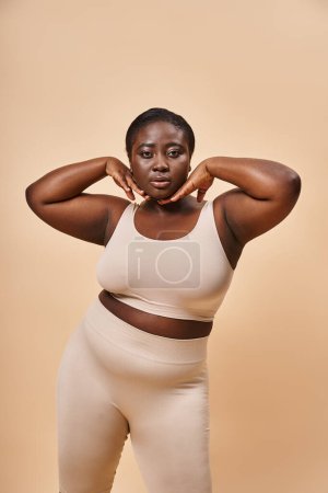 Plus size African American woman in beige underwear posing with hands near face on beige background
