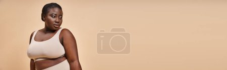 Plus size African American woman in beige undergarments posing on beige background, banner