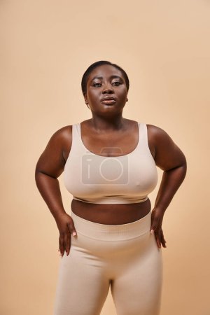Plus size African American woman in beige underwear looking at camera on beige background